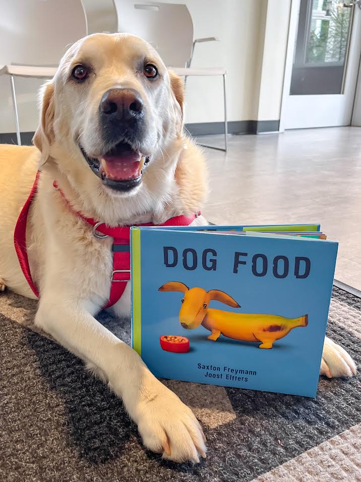 Shelby a therapy dog poses with a children's book called Dog Food during Rescue Readers