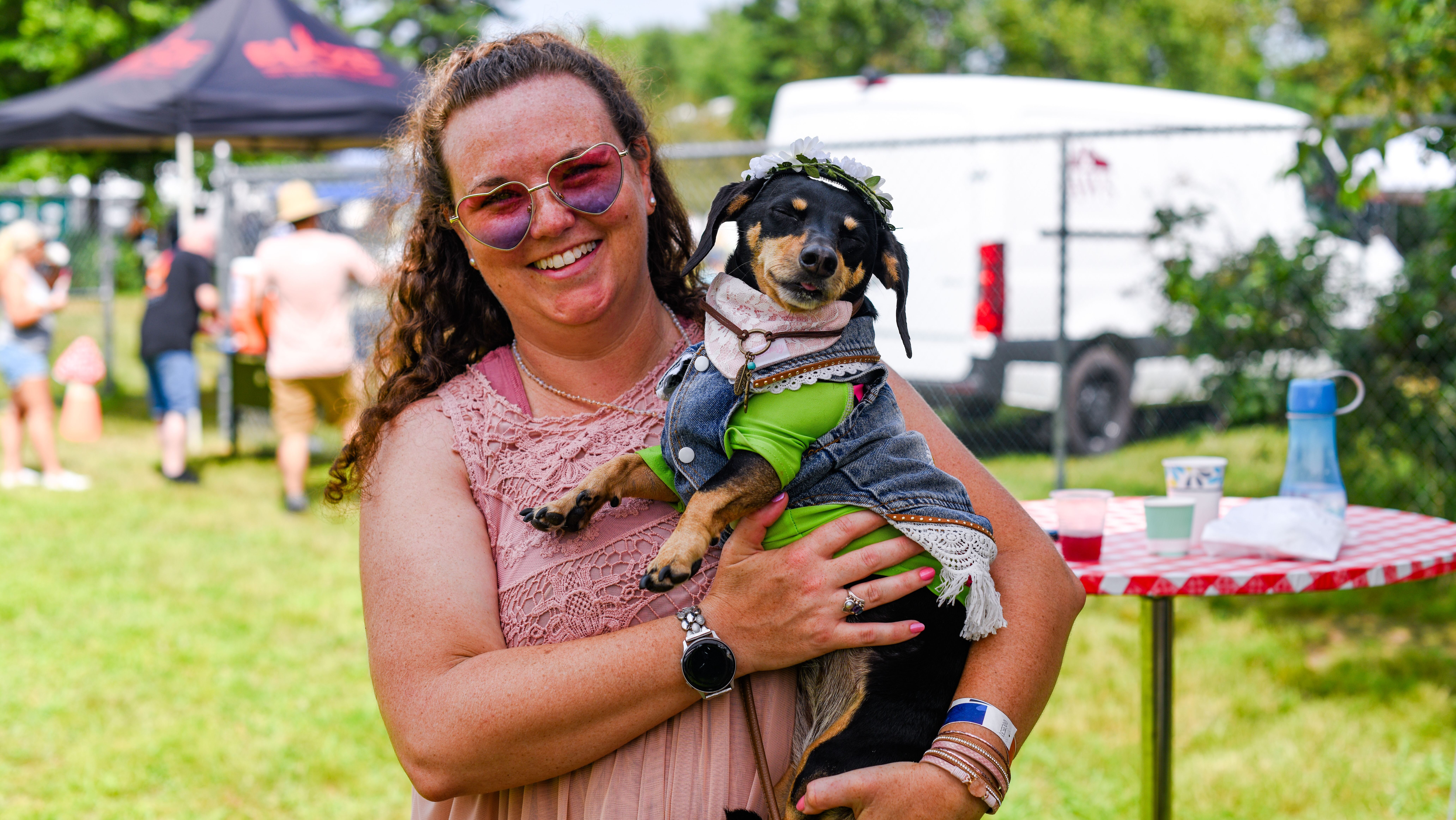 A woman wearing heart-shaped glasses holds her small dog who is dressed in hippie clothes.