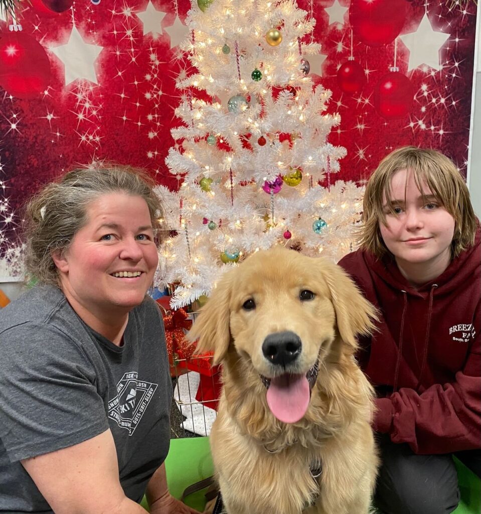 A mother and her child sit next to their dog in front of a Christmas tree and smile.