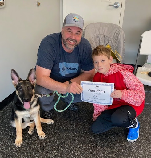 Man and son hold a dog training certificate while puppy sits obediently beside them