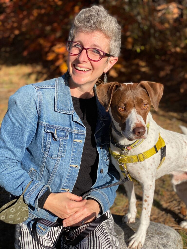 canine trainer in denim jacket poses in the sunshine with brown and white dog with yellow harness