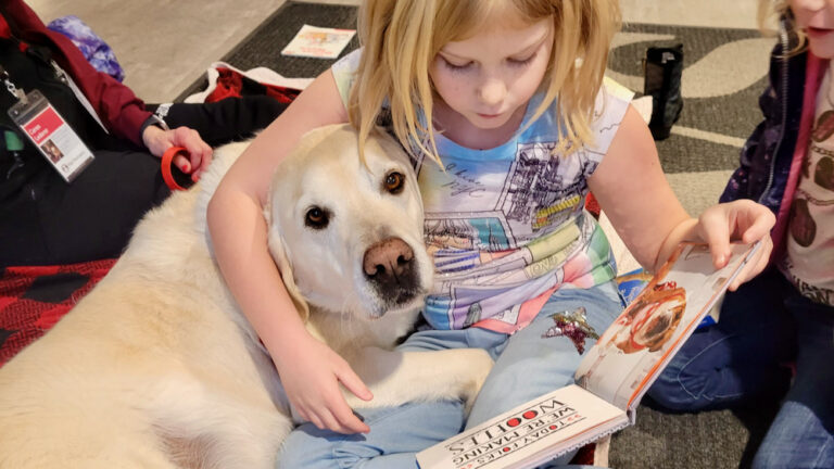 A young girl hugs a dog while she sits on the floor and reads him a book.