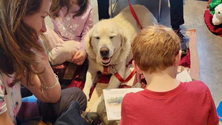 A dog lies on the floor and smiles while being read to by a circle of children.