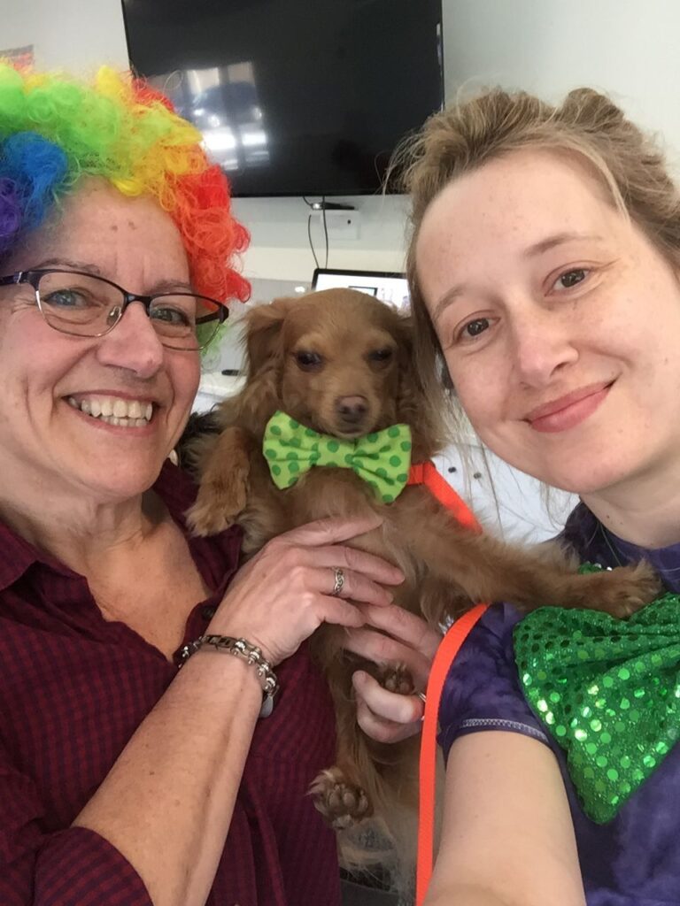 Close-up of two people wearing clown wigs and bowties hug a small dog.