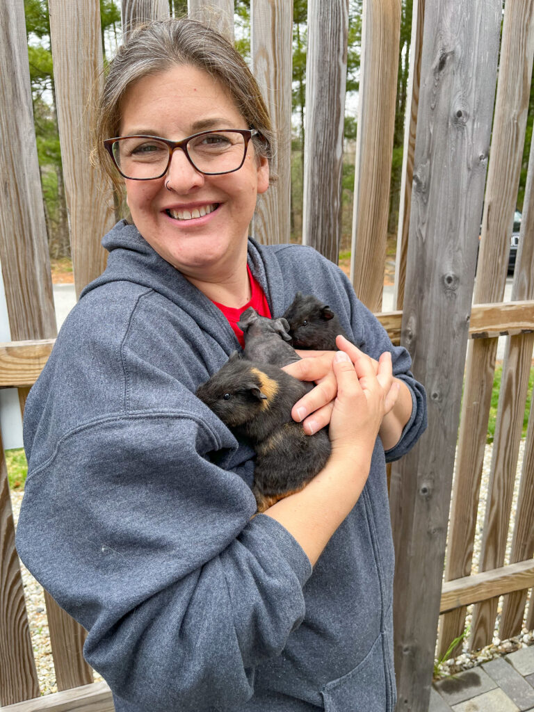 A woman smiles at the camera while holding three baby guinea pigs outside as part of AWS' social-emotional learning program. 