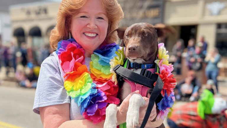 A woman wearing a lei holds a puppy also wearing a lei in a parade.