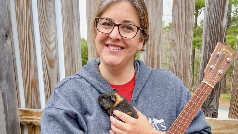A woman smiles while holding a guinea pig and a ukulele in front of a fence.