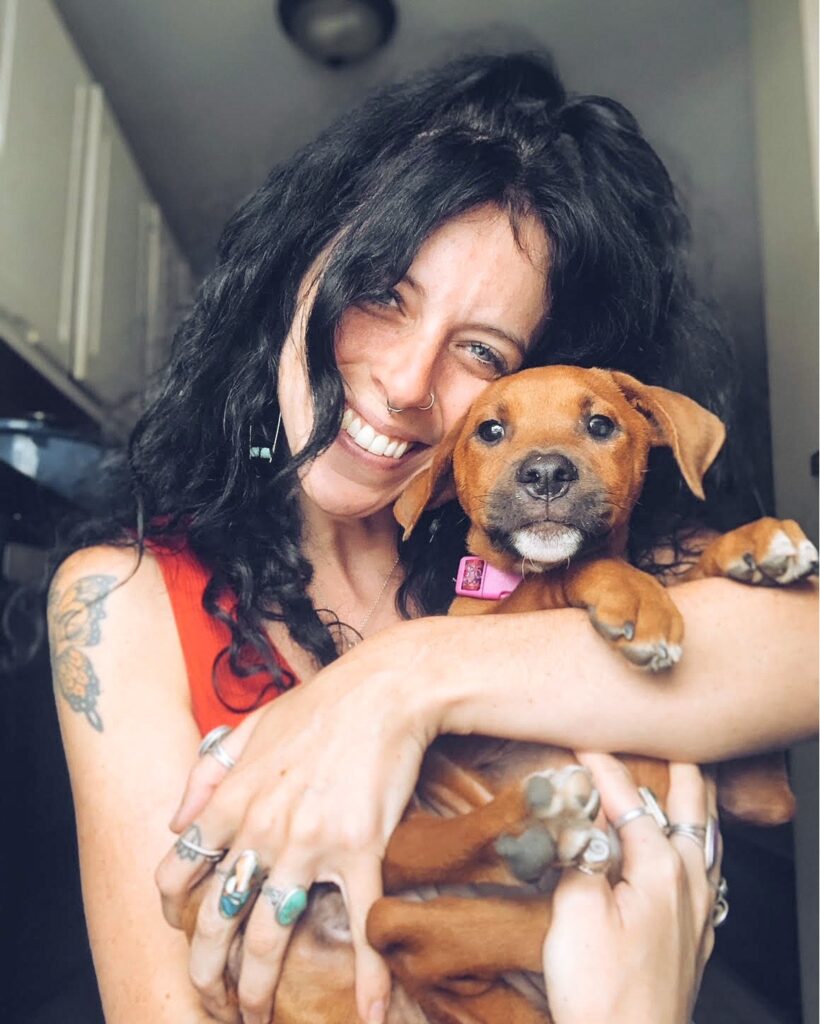 An AWS foster volunteer looks at the camera while holding a brown puppy.