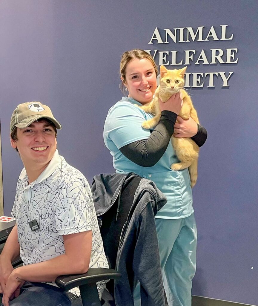 man sitting and a woman veterinarian standing and holding an orange cat welcome new clients to AWS Community Veterinary Clinic