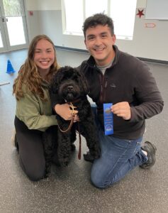 Two graduates of a Canine Good Citizen prep class along with their pup holding up their ribbon.