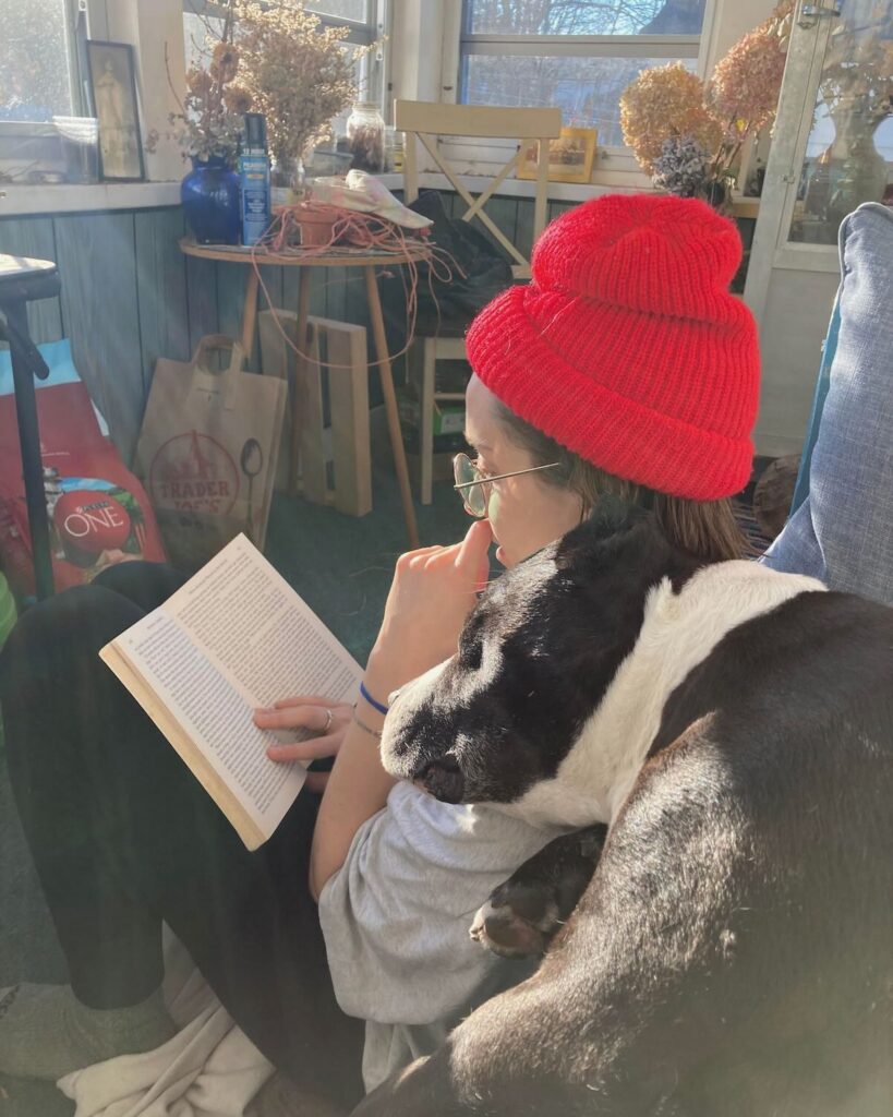 a college aged woman in a red hat sits and studies on a couch while her foster dog cuddles his head on her shoulder
